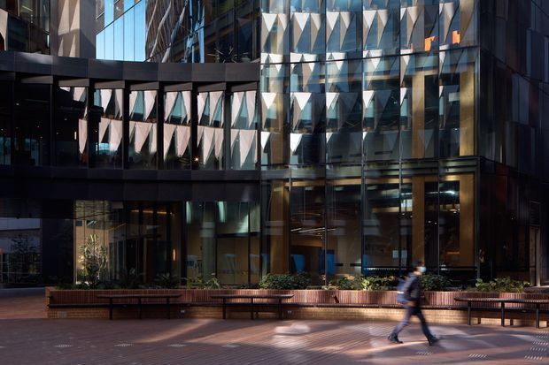 Urban commercial building facade with extensive floor-to-ceiling glazing, dark metal framing, and distinctive triangular geometric sun-shading or patterned facade elements. The image is viewed from a landscaped pedestrian plaza with integrated timber-toned planter seating, benches, and a blurred walking figure. Reflections of surrounding buildings are visible in the glass.