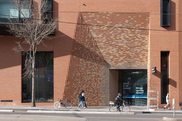 Street-level exterior view of a contemporary urban brick building. The facade uses red brick with a prominent recessed and projecting patterned brick entrance canopy, large dark-framed windows, and a glazed doorway. Cyclists and pedestrians are present along the sidewalk, reinforcing the active city setting.
