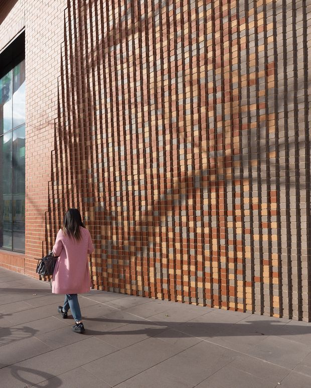 Exterior architectural detail of a contemporary brick building. A large patterned facade combines projecting and recessed red, orange, tan, and dark brown bricks in a vertical grid, beside tall dark-framed glazing. A paved pedestrian walkway and a person provide scale.