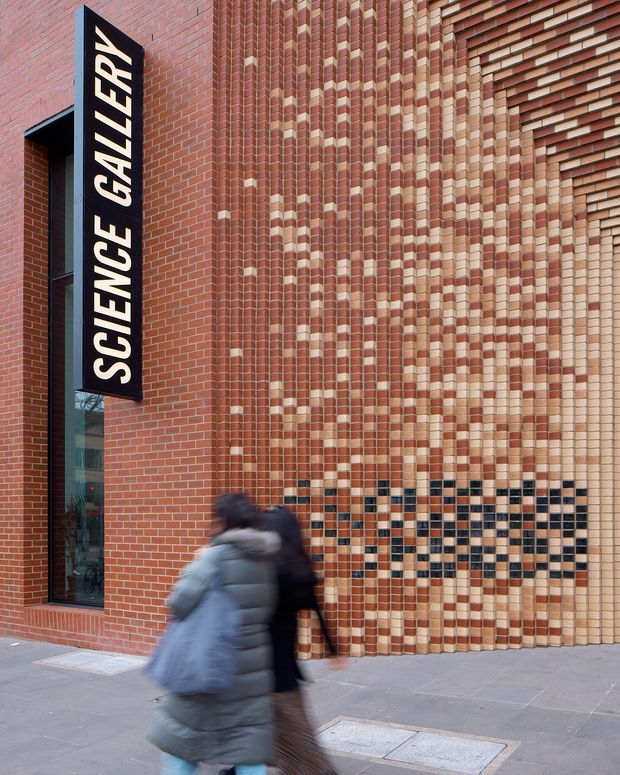 Street-level exterior view of a brick-clad cultural building marked “SCIENCE GALLERY.” The facade combines red brickwork with projecting and recessed patterned brick detailing, including a geometric band of darker blue-black and pale bricks near the base. A tall black vertical sign and a narrow glazed entrance opening are visible, with pedestrians passing in the foreground.