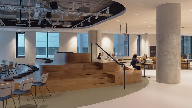 Modern commercial interior with a large tiered timber presentation platform and integrated seating, surrounded by tables, chairs and people. The space features exposed services and ducts within a dark oval ceiling recess, smooth white ceilings, large windows, concrete columns, pendant lighting and polished light flooring. It appears to be a contemporary workplace, educational, hospitality or event space designed for presentations and informal collaboration.
