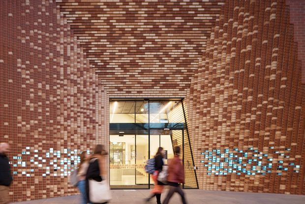 Architectural view of a covered passage or entry portal formed by extensive textured brickwork. The walls and soffit use a dense patterned arrangement of reddish-brown bricks with lighter projecting accents and bands of blue glazed or illuminated brick elements near the base. A glazed entrance with black metal framing and an internal mezzanine is visible at the far end, while blurred pedestrians provide scale. The image has a contemporary yet strongly material-focused civic or commercial character.