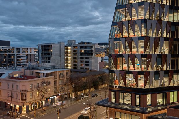 Twilight urban architectural view overlooking a city street, with a prominent contemporary multi-storey building in the foreground. The building features extensive floor-to-ceiling glazing, dark framing, and dramatic repeating triangular facade panels, with illuminated interiors visible inside. Mid-rise apartment and commercial buildings, historic street-front architecture, roads, and street trees surround it.
