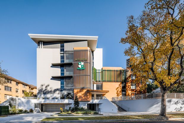 Exterior view of a contemporary multi-storey institutional or commercial building with strong geometric forms, white rendered walls, dark grey upper cladding, projecting flat rooflines, vertical fins, balconies, and a glazed enclosed bridge. The building sits on a landscaped urban site beside mature trees, with a recessed ground-level entry and garage-like openings.