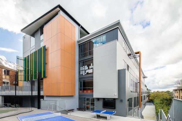 Exterior view of a contemporary multi-storey building with a bold orange panel-clad vertical volume, white and grey facade sections, extensive glazing, projecting roof planes, and colourful vertical screening. The foreground includes an outdoor recreational court or play area, while neighbouring urban buildings and trees are visible in the background.