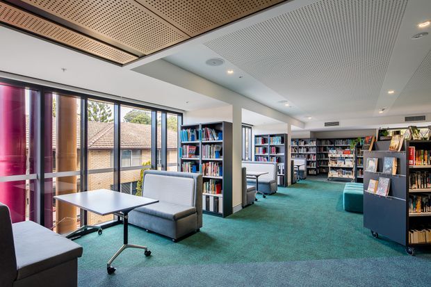 A bright, contemporary library or study space with floor-to-ceiling glazing, extensive mobile book shelving, individual study tables, upholstered lounge chairs, and teal carpet. The ceiling combines perforated timber-look acoustic panels with a white linear acoustic treatment and recessed lighting. A brick building is visible through the windows.