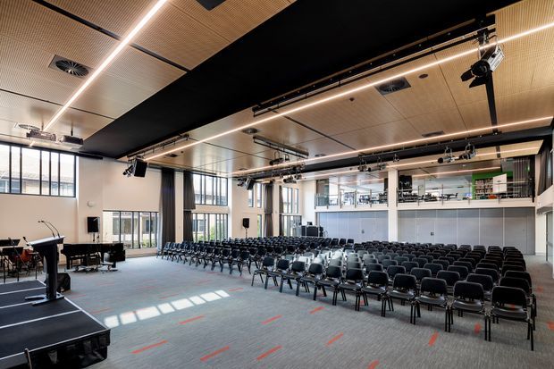 A large contemporary auditorium or multipurpose performance and event hall with rows of black chairs facing a stage and grand piano. The space features high ceilings with acoustic timber slatted panels, black ceiling sections, linear strip lighting, theatrical rigging, large windows, and an upper-level balcony or viewing area.