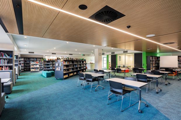A bright contemporary library or learning commons with extensive bookshelves, rows of movable tables and chairs, turquoise carpet, and green-accented walls. The ceiling features warm timber acoustic paneling with integrated linear lighting, recessed services, and ventilation. A presentation screen and open collaborative study area are visible.
