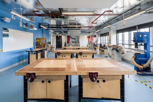A bright, spacious industrial-style woodworking and fabrication workshop with multiple large timber workbenches, bench vises, blue-painted machinery, dust extraction hoses, shelving, a whiteboard, and exposed ceiling services. The room has blue walls and flooring, extensive overhead ducting and pipework, and large windows along one side.