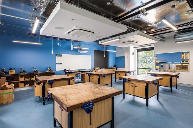 A bright, contemporary workshop classroom or maker space with multiple heavy-duty timber workbenches, blue-painted walls, exposed services and reflective insulation at the ceiling, suspended linear lighting, wall-mounted tools, and a large whiteboard. The room has a practical educational or fabrication setting with generous daylight through a window.