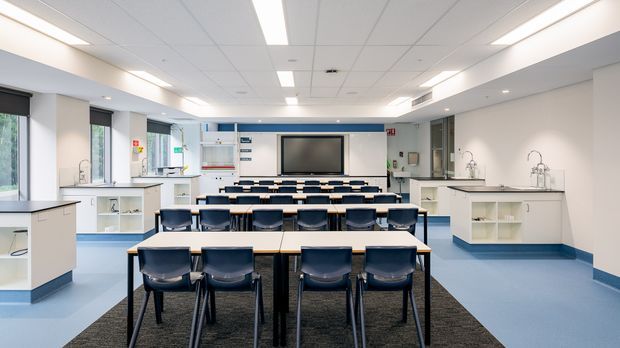 Bright contemporary commercial teaching and training space with rows of tables and dark chairs facing a wall-mounted display. The room includes white built-in cabinetry, multiple sinks and work counters along both sides, blue resilient flooring, dark carpeted zones, extensive glazing, and a suspended acoustic tile ceiling with integrated linear lighting.