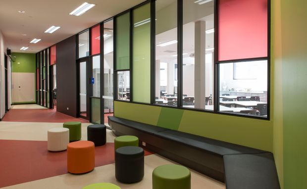 A contemporary commercial interior corridor or communal breakout area with black-framed glazed partitions, colourful translucent and opaque wall panels, integrated dark bench seating, and scattered cylindrical ottomans. The space has a bold red, cream, green, and charcoal palette, fluorescent linear ceiling lights, and an open office visible through the glazing.