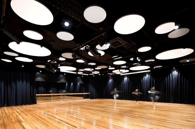 A large, dark-lined studio or performance space with a highly polished timber floor, full-height black curtains, mirrored wall panels, extensive theatrical lighting and rigging, and numerous large circular illuminated ceiling panels. Several blurred people are moving across the open floor, suggesting a dance or rehearsal studio.