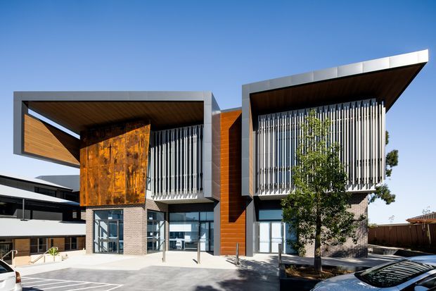 Front exterior view of a contemporary multi-storey building with two projecting box-like upper volumes, broad flat roof overhangs, vertical metal screening, warm timber cladding, exposed weathered steel feature panels, brickwork, and extensive glazing. The building fronts a paved forecourt with mature trees and parked vehicles at the edges.