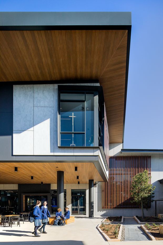 Exterior architectural photograph of a contemporary multi-storey institutional or commercial building. The facade combines dark metal framing, light grey panel cladding, extensive glazing, warm timber soffits, cylindrical columns, and a vertical timber screen. A covered entrance and paved forecourt are visible, with people providing scale.