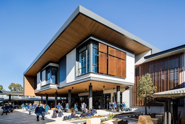 Contemporary multi-storey building with a dramatic cantilevered form, broad pitched roof planes, exposed timber soffits, white cladding, dark structural columns, and extensive glazing. The busy forecourt and adjacent slatted facade suggest a public or commercial campus setting.
