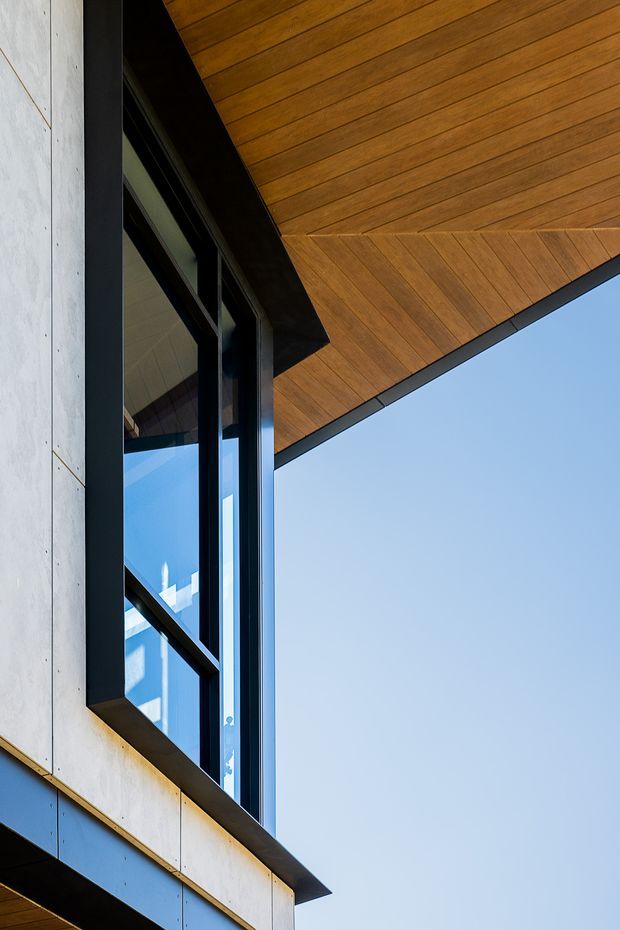 Close-up exterior architectural view showing a contemporary facade with pale concrete or fibre-cement wall panels, dark metal window framing, expansive glazing, and a warm timber-lined projecting soffit. The composition emphasizes clean geometric lines and the junction between the facade, roof overhang, and sky.
