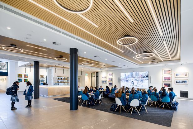 A bright, contemporary commercial interior resembling a school or institutional common area, with a café or reception counter, clustered tables and chairs, black structural columns, framed artwork, and a large wall-mounted display. The space features a striking linear timber slatted ceiling with integrated circular and linear lighting, contrasting with white ceiling panels and pale flooring.