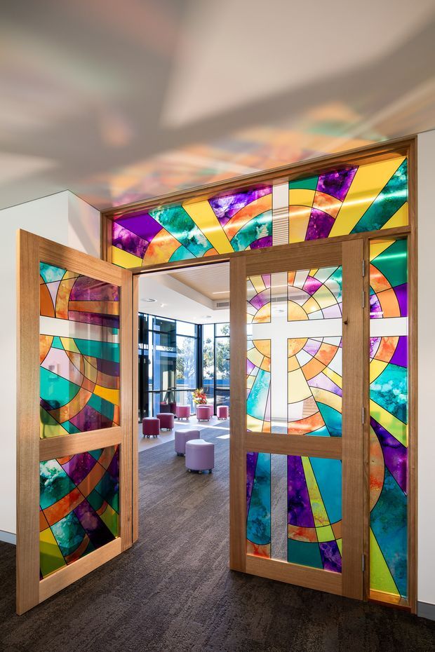 A bright commercial interior entrance featuring tall timber-framed double doors and transom panels filled with vivid abstract stained glass in purple, teal, turquoise, yellow, orange, and cream. The doors open into a contemporary lounge or reception area with large glazing, carpet flooring, and small pink upholstered stools. Reflections of the colourful glass cast patterns across the ceiling.