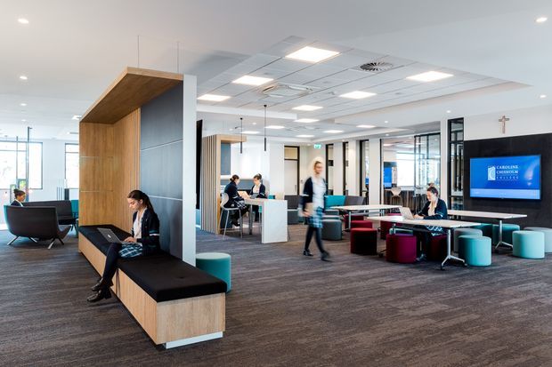 A bright contemporary commercial interior, likely a school or institutional learning commons, with open-plan work and collaboration areas. The space features carpeted flooring, white ceilings with recessed lighting and services, timber-lined built-in seating, black-framed glazed partitions, desks, stools, lounge seating, and a large wall-mounted display.