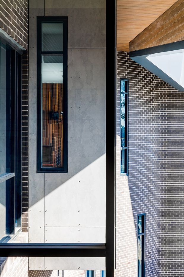 Vertical architectural exterior detail showing a contemporary multi-level building. The facade combines dark brickwork, large grey concrete or fibre-cement panels, black-framed narrow windows, deep black structural reveals, and warm timber soffit cladding. Strong sunlight creates sharp geometric shadows across the facade, emphasizing the building’s angular composition and recessed openings.