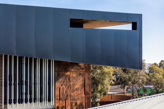 Exterior architectural photograph of a contemporary building with a bold cantilevered upper volume clad in dark blue-grey panels. A long horizontal opening reveals a warm timber soffit, while the lower facade combines vertical black-and-white fins with weathered rust-toned metal panels. A corrugated metal roof and suburban trees are visible in the foreground and background.