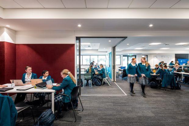 A contemporary open-plan educational or commercial interior with students and staff working at shared tables. The space features carpeted flooring, white ceilings with recessed lighting, a deep red acoustic feature wall, extensive internal glazing, and collaborative work areas furnished with desks and chairs.
