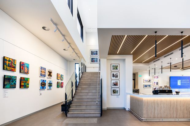 Bright contemporary gallery or commercial reception interior with a central staircase, white display walls hung with colourful artwork, framed displays along the stair landing, and a timber-fronted reception counter. The space has a high open ceiling, linear timber slatted ceiling feature, integrated track and pendant lighting, polished concrete or stone-look flooring, and extensive glazing near the stair.