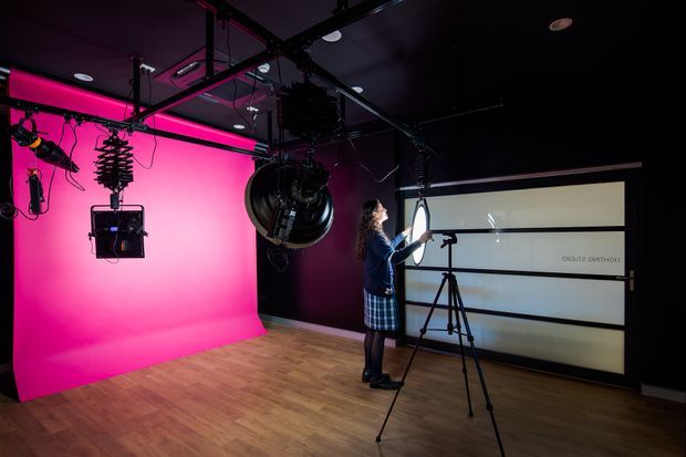 A professional photography or film studio with a vivid pink cyclorama backdrop, extensive overhead lighting and rigging, a tripod-mounted camera, and a person working near a large illuminated display or backdrop. The room has dark walls and ceiling with a timber-look floor, creating a dramatic production environment.