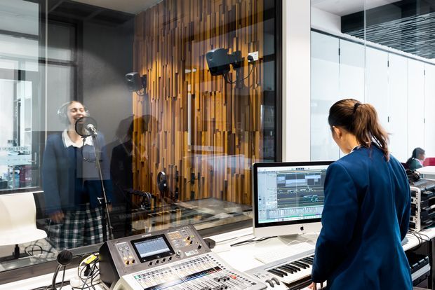 A contemporary professional recording studio viewed through a glass partition. In the foreground, a sound engineer operates a large audio mixing console beside a computer workstation and keyboard. Behind the glazing, a vocalist performs into a microphone inside a booth with acoustic treatment, including a prominent vertical timber slat feature wall. The space has bright white surfaces, extensive glazing, and dark linear ceiling detailing.