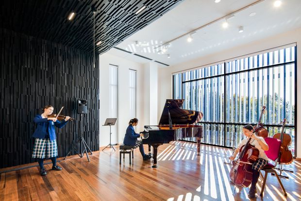 A bright contemporary music performance and rehearsal space with polished timber flooring, a glossy black grand piano, violinist, pianist and cellist, and a dramatic black acoustic slat wall and ceiling. Full-height glazed windows with vertical fins bring in strong daylight and cast striped shadows across the floor.