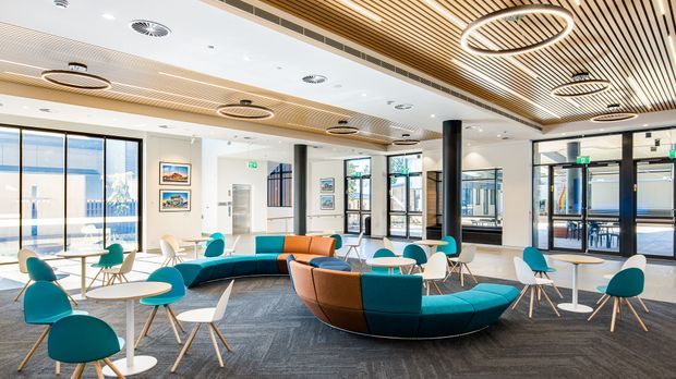 A bright contemporary commercial lounge or reception area with extensive floor-to-ceiling glazing, teal and tan curved modular seating, small round tables, and scattered teal-and-white chairs. The space features a dark carpeted floor, white walls, black structural columns, a slatted timber-look ceiling with integrated circular pendant lights, and visible entry doors and windows.