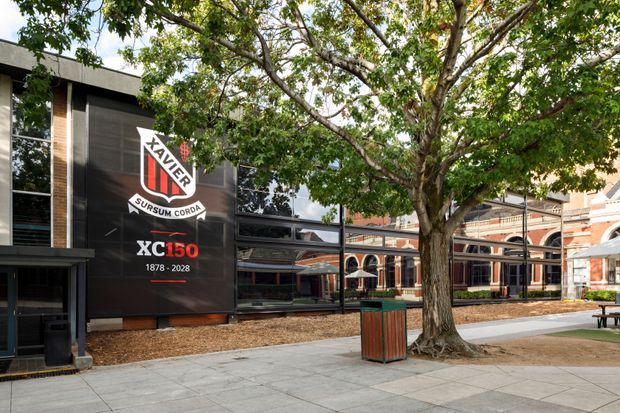 A tree-lined paved courtyard or campus walkway beside a contemporary dark-clad building. The facade features large reflective windows and a prominent Xavier College anniversary banner, while an older brick building with arched colonnades is visible beyond the courtyard. Outdoor seating and a timber-and-metal rubbish bin occupy the landscaped pedestrian area.