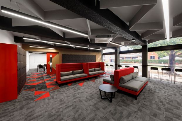 A contemporary commercial interior, likely a collaborative lounge or reception area, with red upholstered booth seating, grey lounge chairs, black tables, extensive glazing, and a dramatic exposed geometric ceiling. The space uses a bold red, black, and charcoal palette with patterned carpet and linear strip lighting.