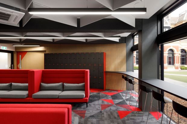 A contemporary commercial interior, likely a student or workplace lounge, with vivid red upholstered booth seating, a long window-side bar counter with stools, dark locker-style cabinetry, and a geometric red, grey, and black carpet. Large black-framed windows overlook a heritage-style brick building. The ceiling features sculptural white acoustic panels and linear pendant lighting.