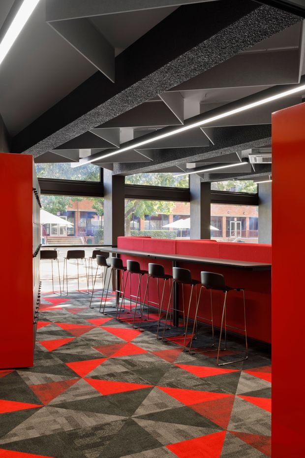 A bold contemporary commercial interior, likely a café, hospitality venue, or collaborative workplace, with a long red upholstered banquette and bar-height counter lined with black stools. Floor-to-ceiling windows bring in daylight, while the ceiling features dramatic geometric acoustic panels and linear strip lighting. The carpet uses a striking geometric pattern in red, charcoal, grey, and beige, complemented by large red partitions or joinery elements.