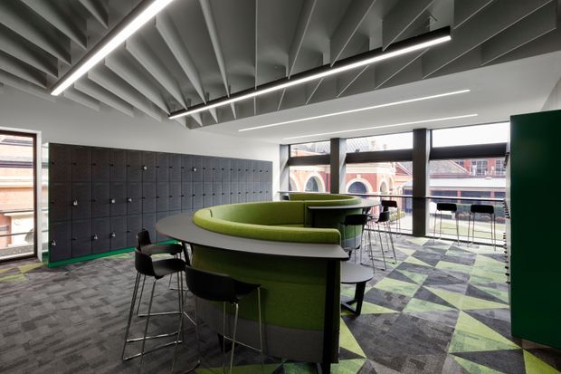 A contemporary commercial interior featuring a collaborative breakout and work area. Curved green upholstered booth seating wraps around a central high table, with additional stools and chairs. The space has a dramatic exposed ribbed ceiling, linear pendant lighting, dark locker-style wall storage, floor-to-ceiling windows, and geometric grey, green, and purple carpet tiles. Historic-looking buildings are visible outside the glazing.