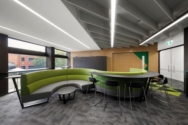 A contemporary commercial lounge and collaborative breakout area with curved lime-green upholstered banquette seating surrounding a large oval communal table. The space has dark patterned carpet, timber wall panelling, grey exposed ceiling beams, black-framed glazing, linear LED lighting, lockers, and a bold green geometric wall feature. A glass entry door and exit signage are visible at the right.