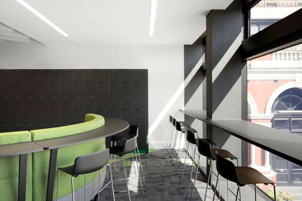 A bright contemporary commercial interior, likely a staff breakout or shared workplace lounge. The space features a long curved lime-green upholstered banquette around a dark rounded table, black and grey bar stools, a wall of dark lockers, charcoal structural columns, large windows, and a high counter along the glazing. Linear recessed ceiling lights and patterned carpet reinforce the modern office fit-out. A historic-looking building is visible outside through the windows.