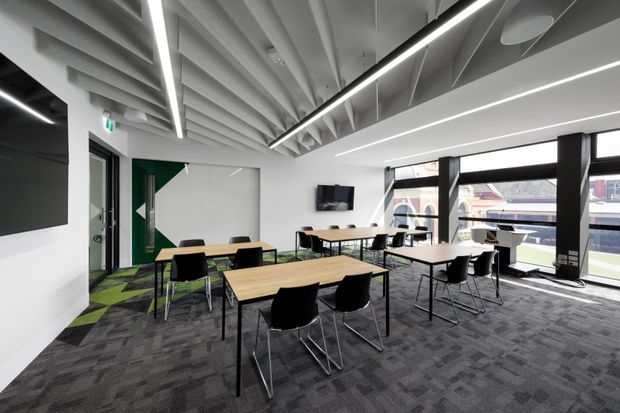 Contemporary open-plan commercial meeting and collaboration space with multiple light timber tables, black chairs, dark patterned carpet, large windows, and exposed ceiling structure. Linear LED lighting runs through the high ceiling, while a green geometric wall feature and wall-mounted display add visual accents.