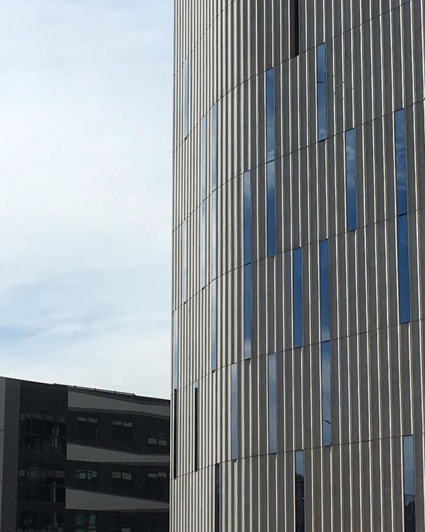 Exterior architectural photograph of a tall contemporary building facade with closely spaced vertical fins and narrow blue-tinted window strips arranged in a rhythmic pattern. A darker multi-storey building is partially visible in the background beneath an overcast sky.