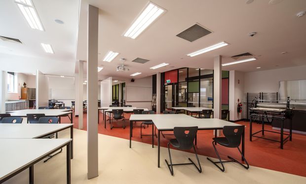 A bright, open commercial interior arranged as a flexible dining, meeting, or educational space. Multiple white rectangular tables and black chairs sit across a large room with red carpet zones and pale flooring. The space includes exposed structural columns, extensive fluorescent ceiling lighting, acoustic ceiling panels, glazed internal partitions, a projector, and what appears to be a games or recreation area at the rear. The overall fit-out is practical, institutional, and contemporary.