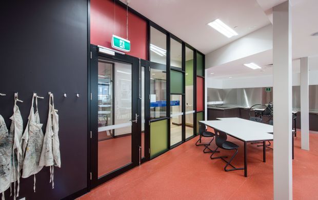 A contemporary commercial interior with an open meeting or breakout area, white rectangular tables, black chairs, and a kitchenette or work counter at the rear. Black-framed glazed partitions with translucent and coloured panels divide the space, while red flooring and a dark feature wall create a bold, functional workplace setting. Fluorescent ceiling lighting and exit signage are visible.