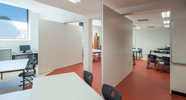 Bright contemporary commercial interior, likely a flexible classroom, training room, or office workspace. The open-plan area contains white tables, black chairs, computer workstations, freestanding white partition walls, large windows with blinds, and flush-mounted fluorescent ceiling lighting. A red terracotta-toned resilient floor contrasts with white walls and furniture.