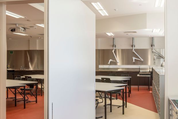 A bright commercial teaching kitchen or culinary classroom viewed through an open doorway. Multiple stainless-steel workstations, sinks, wall-mounted extraction or service fixtures, tables, chairs, and overhead fluorescent lighting are arranged across a spacious room. The floor combines red and pale cream finishes, with white walls and stainless-steel backsplashes and equipment.
