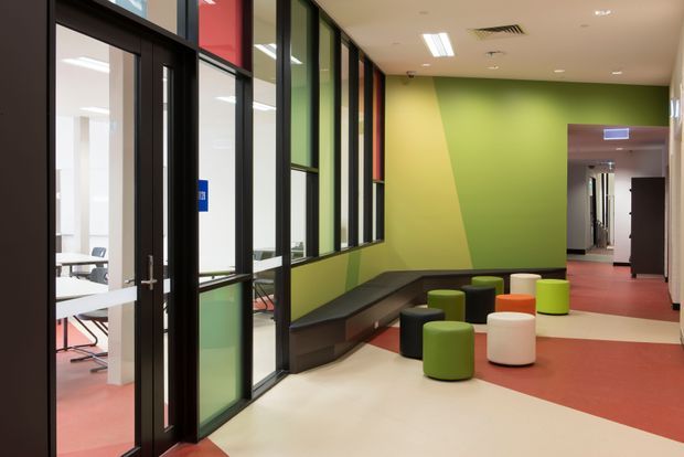 Interior view of a colorful institutional or commercial corridor and informal breakout seating area. A lime-green feature wall runs alongside a built-in dark bench, with assorted round ottomans in green, black, orange, white, and yellow. Black-framed glazed partitions reveal a classroom or meeting room on the left, while the corridor continues through an opening at the rear. The space has a contemporary, functional fit-out with red and cream flooring and fluorescent ceiling lighting.