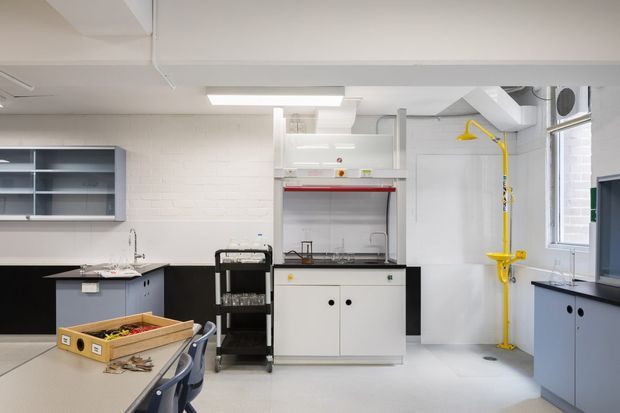 Bright institutional laboratory or workshop interior with white painted brick walls, exposed ceiling services, extensive dark benchtops, blue-grey cabinetry, open shelving, a central white laboratory work cabinet with red trim, sinks, storage, and a prominent yellow emergency shower and eyewash station. Worktables and stools occupy the foreground.