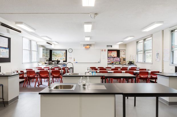 Bright, spacious classroom or teaching laboratory with rows of red chairs and tables, a large central black workbench with integrated stainless-steel sink, whiteboard, display boards, and extensive windows. The room has a clean, functional institutional fit-out with white walls and ceiling, grey flooring, and abundant fluorescent lighting.
