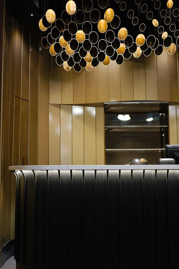 A dramatic, luxurious reception counter or bar-like commercial interior. The foreground features a dark vertically ribbed counter front with a slim light-coloured countertop. Behind it are warm bronze-gold vertical wall panels and reflective dark cabinetry or a service opening. A striking suspended installation of overlapping circular rings and glowing amber spherical elements dominates the ceiling, creating a sculptural focal point. The space is dimly lit with warm, high-contrast illumination and polished reflective surfaces.