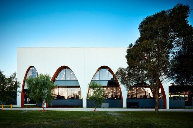 Exterior architectural photograph of a contemporary low-rise commercial building with a clean white facade, repeated large arched glazed openings, and dark red-brown structural arch frames. Interior activity and lighting are visible through the windows. Mature trees, lawn, and a paved frontage sit in front of the building under a clear blue sky.